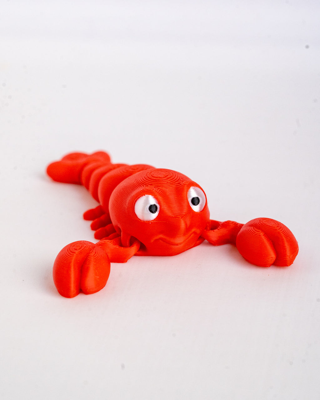 A bright red lobster-shaped 3D-printed toy with large white eyes and black pupils, lying on a plain white surface.