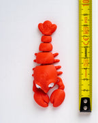 red plastic lobster figurine with white eyes, shown vertically next to a yellow tape measure on a white background.