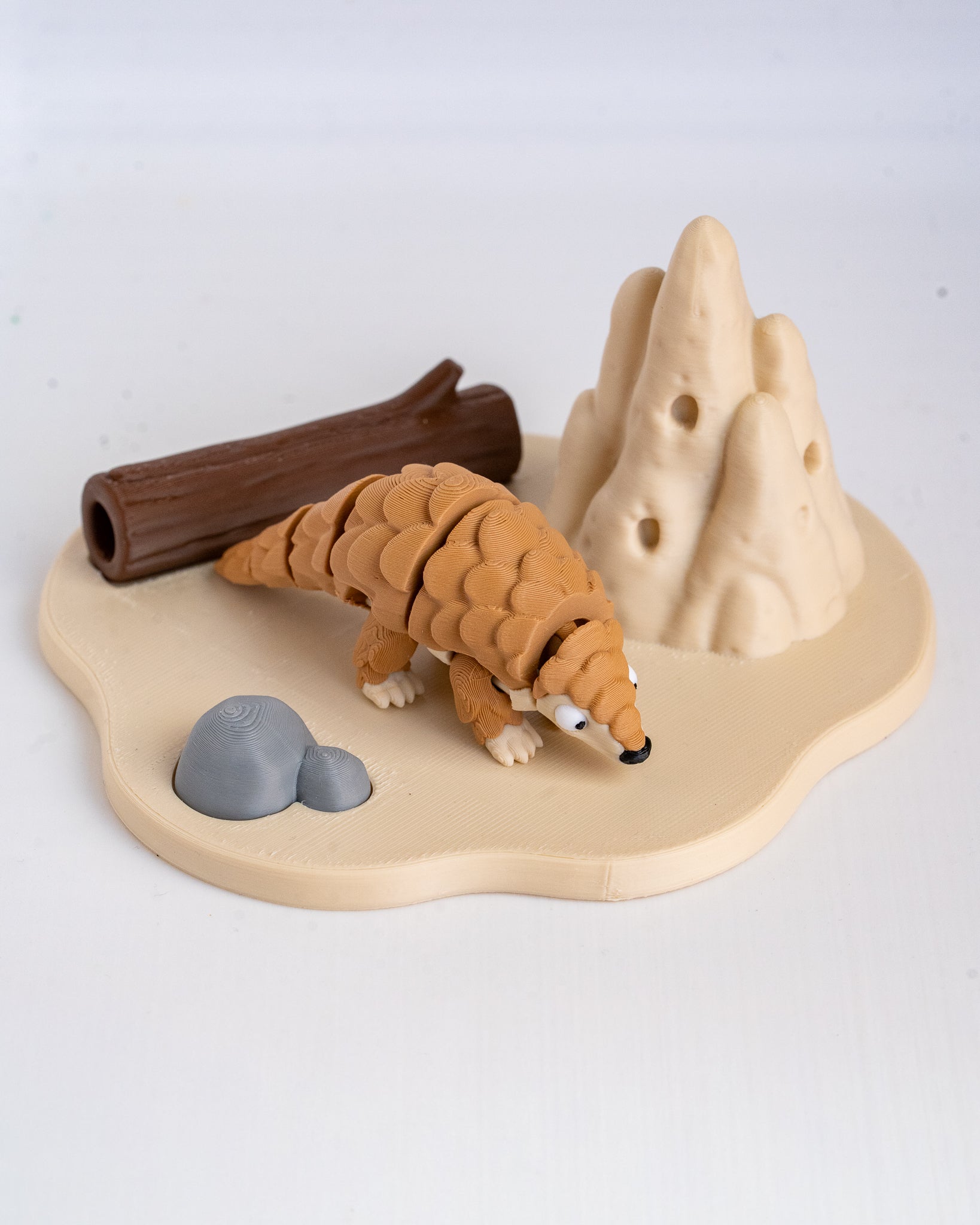 A small animal figurine of a pangolin on a beige base with a brown log, gray rocks, and beige hills, photographed on a white background.