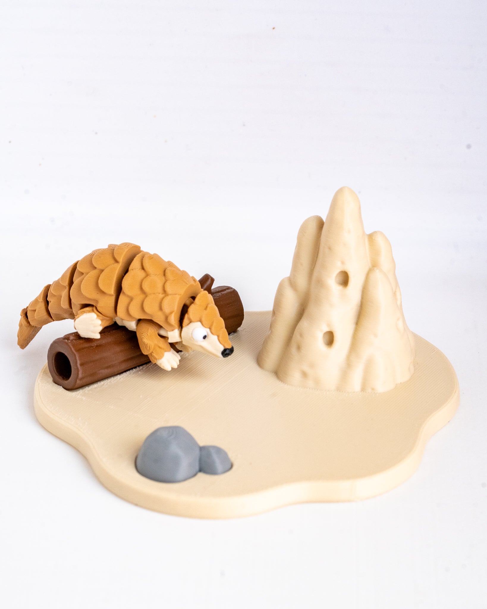 A small animal figurine of a pangolin on a beige base with a brown log, gray rocks, and beige hills, photographed on a white background.