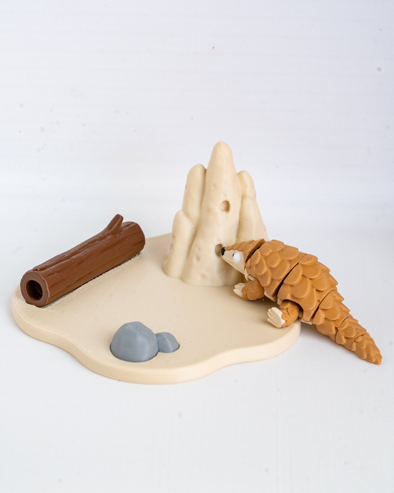 A small animal toy set featuring a brown pangolin figure crawling on a beige base with a beige rock structure, a brown log, and gray rocks, photographed on a white background.