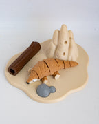 A small animal figurine of a pangolin on a beige base with a brown log, gray rocks, and beige hills, photographed on a white background.
