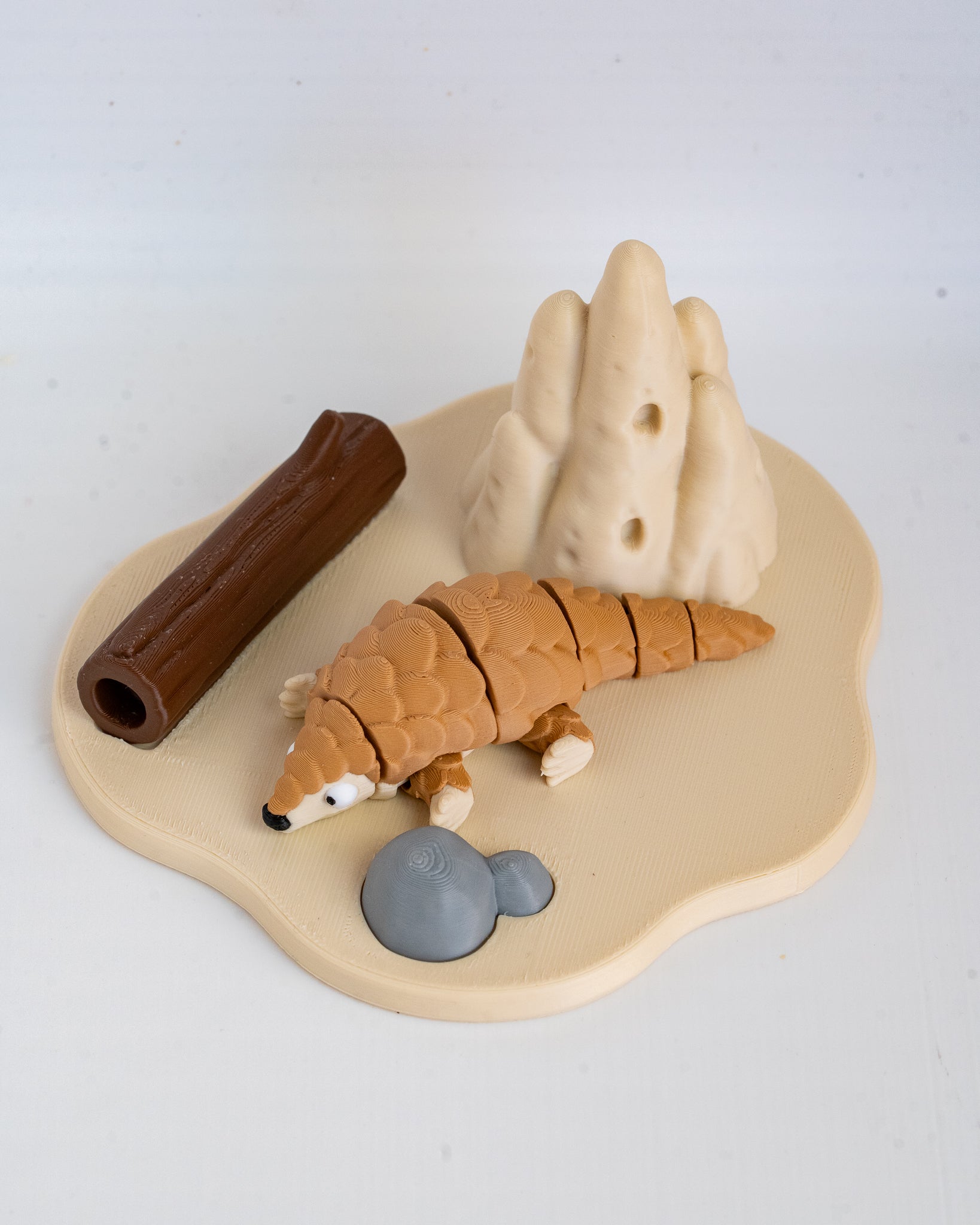 A small animal figurine of a pangolin on a beige base with a brown log, gray rocks, and beige hills, photographed on a white background.