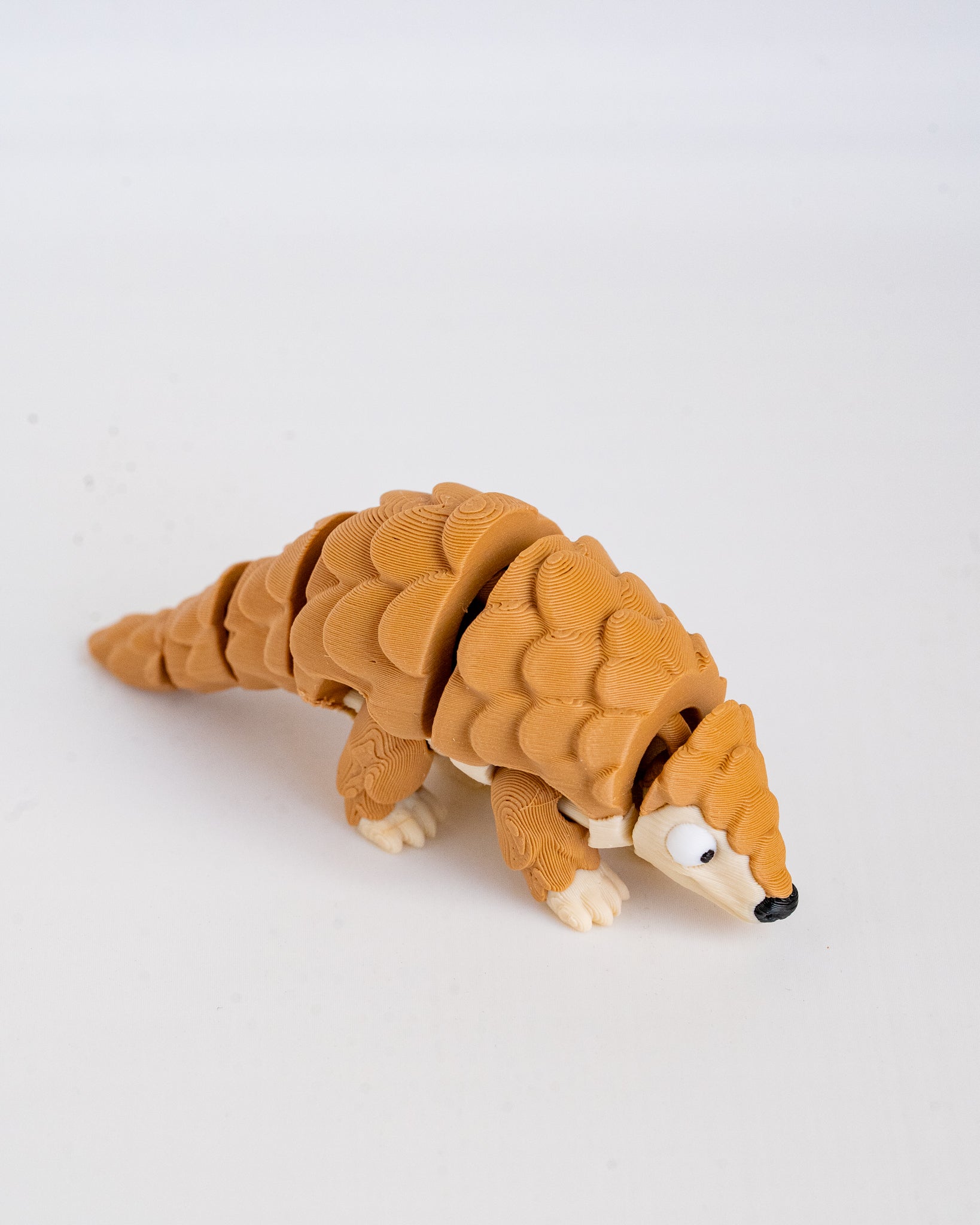 A small articulated pangolin toy figure in a warm tan-brown color with cream-colored feet and snout, photographed on a plain light background.