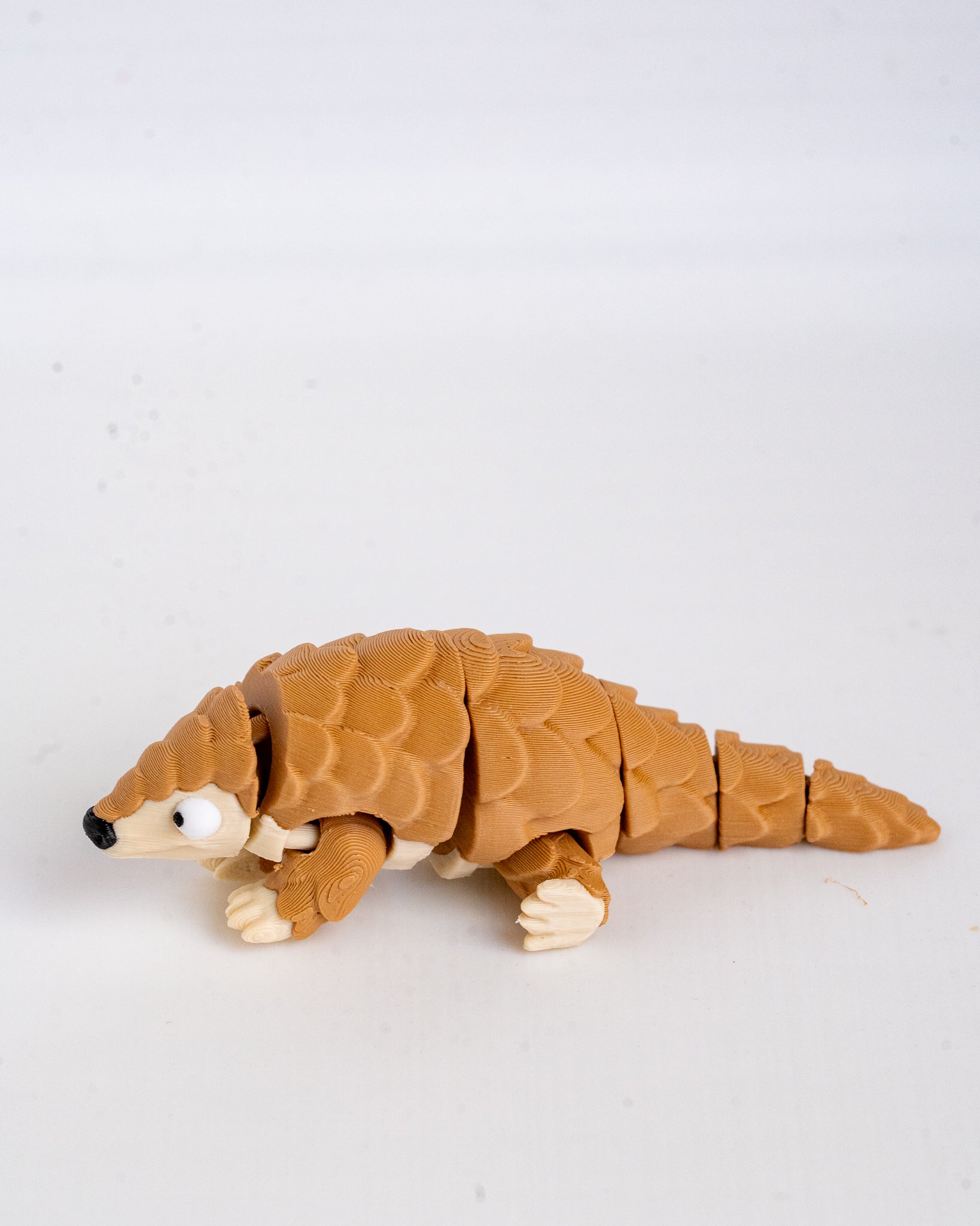 A small articulated pangolin toy figure with textured brown armor plates, cream-colored claws and face details, and a black nose, photographed on a plain light background.