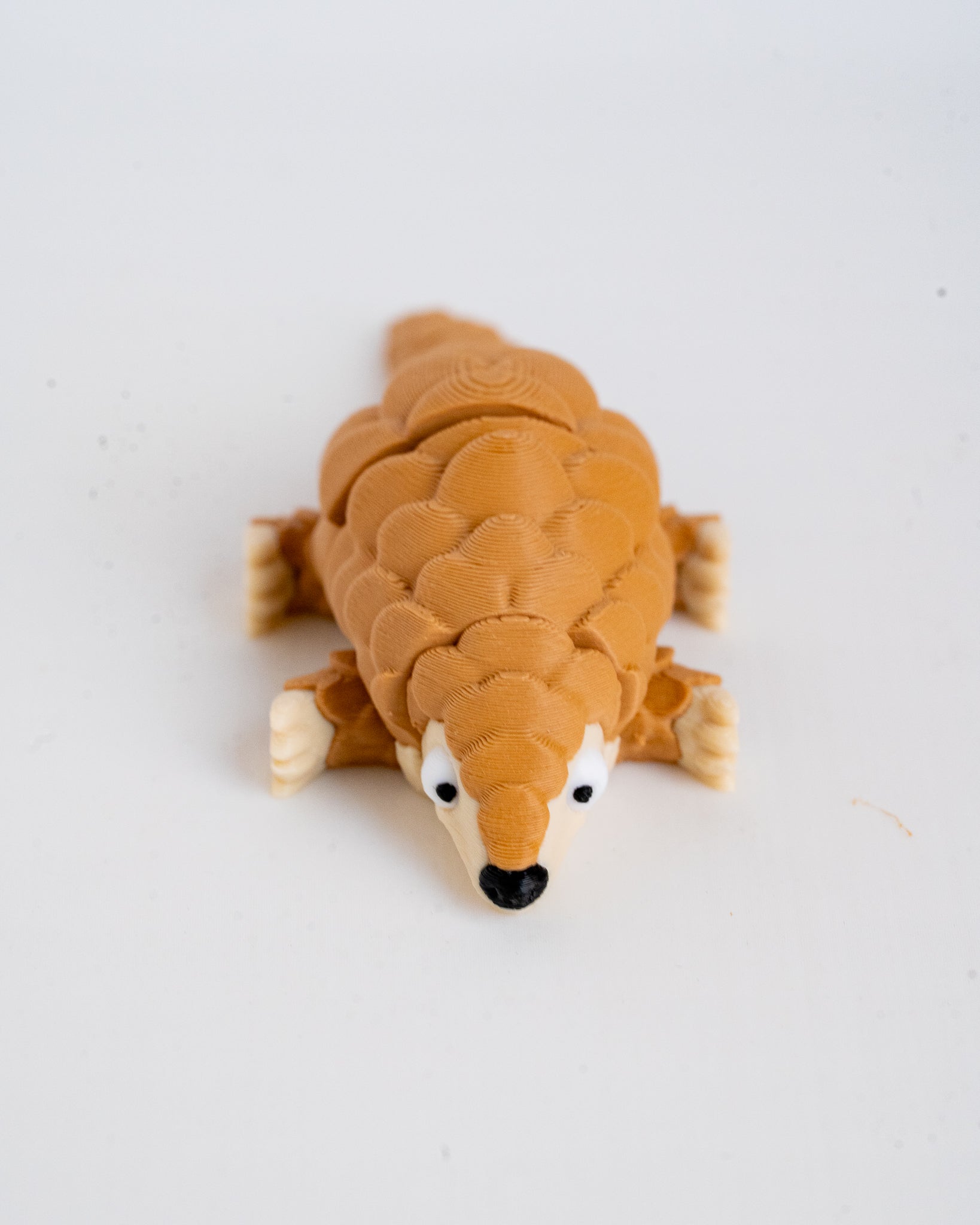 A small pangolin figurine or toy with textured brown scales, cream-colored feet and snout, and a black nose, photographed on a plain light background.