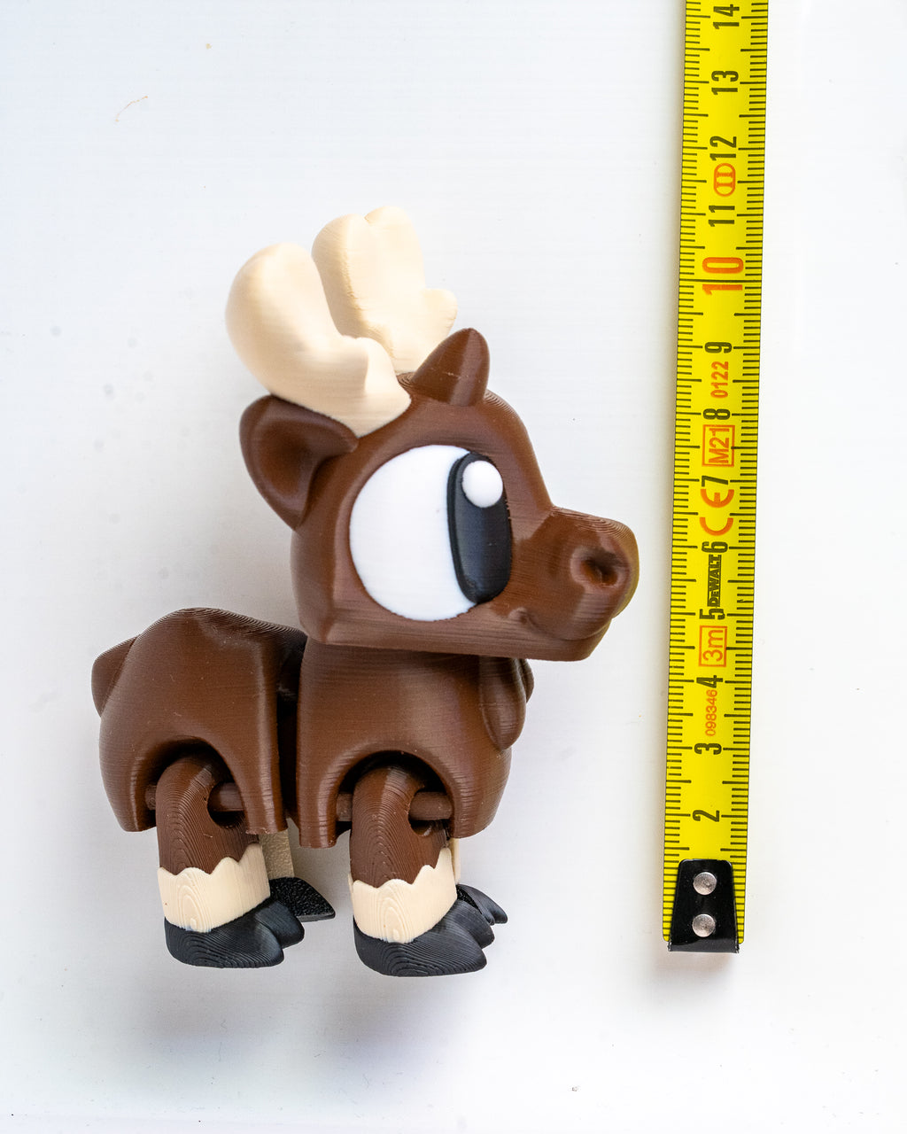 A small brown moose figurine with large white and black eyes, cream-colored antlers and lower legs, standing next to a yellow measuring tape on a white background.