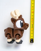 A small brown moose figurine with large white and black eyes, cream-colored antlers and lower legs, standing next to a yellow measuring tape on a white background.