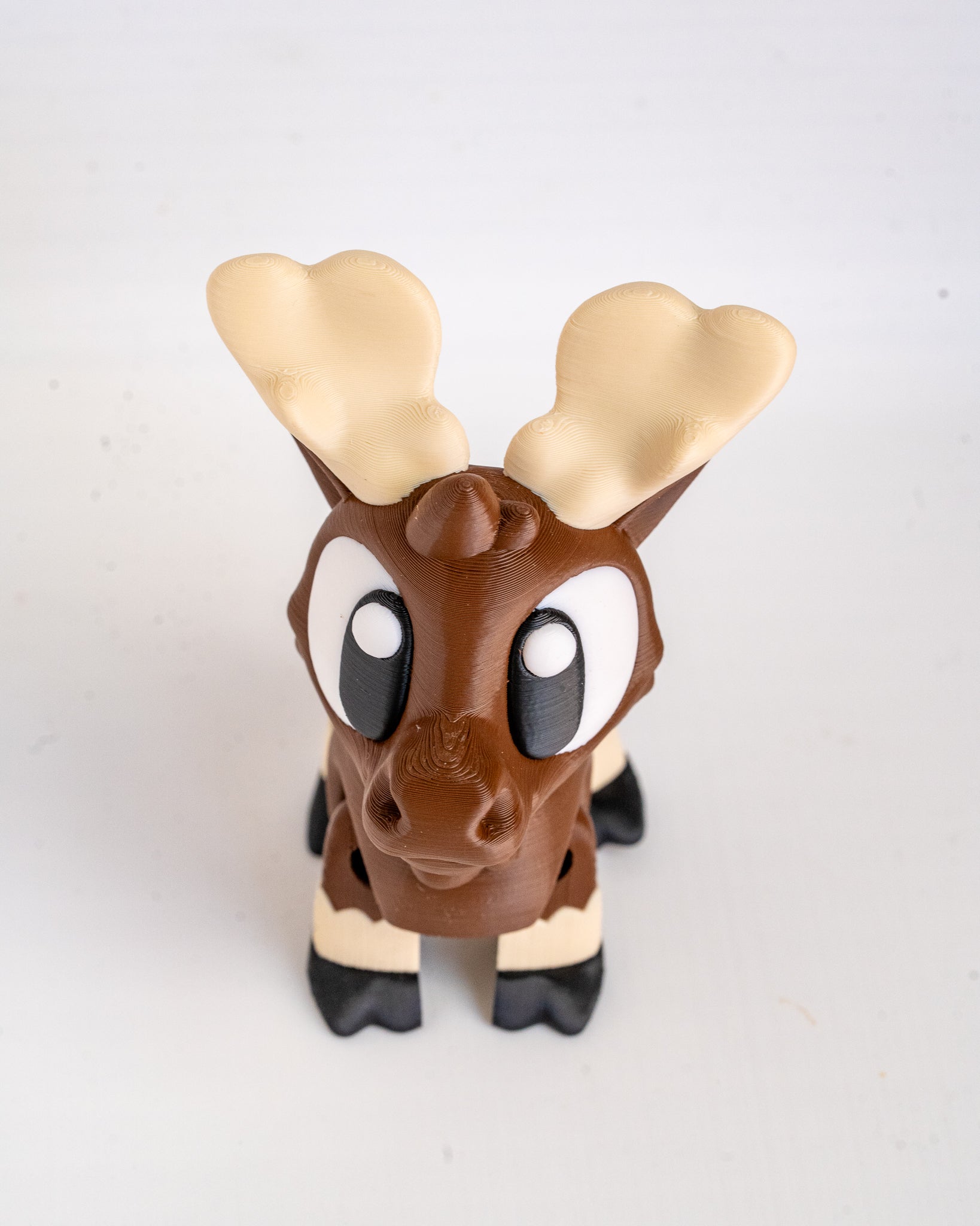A small moose figurine with brown body, cream-colored antlers and lower legs, and black hooves, photographed against a plain light background.