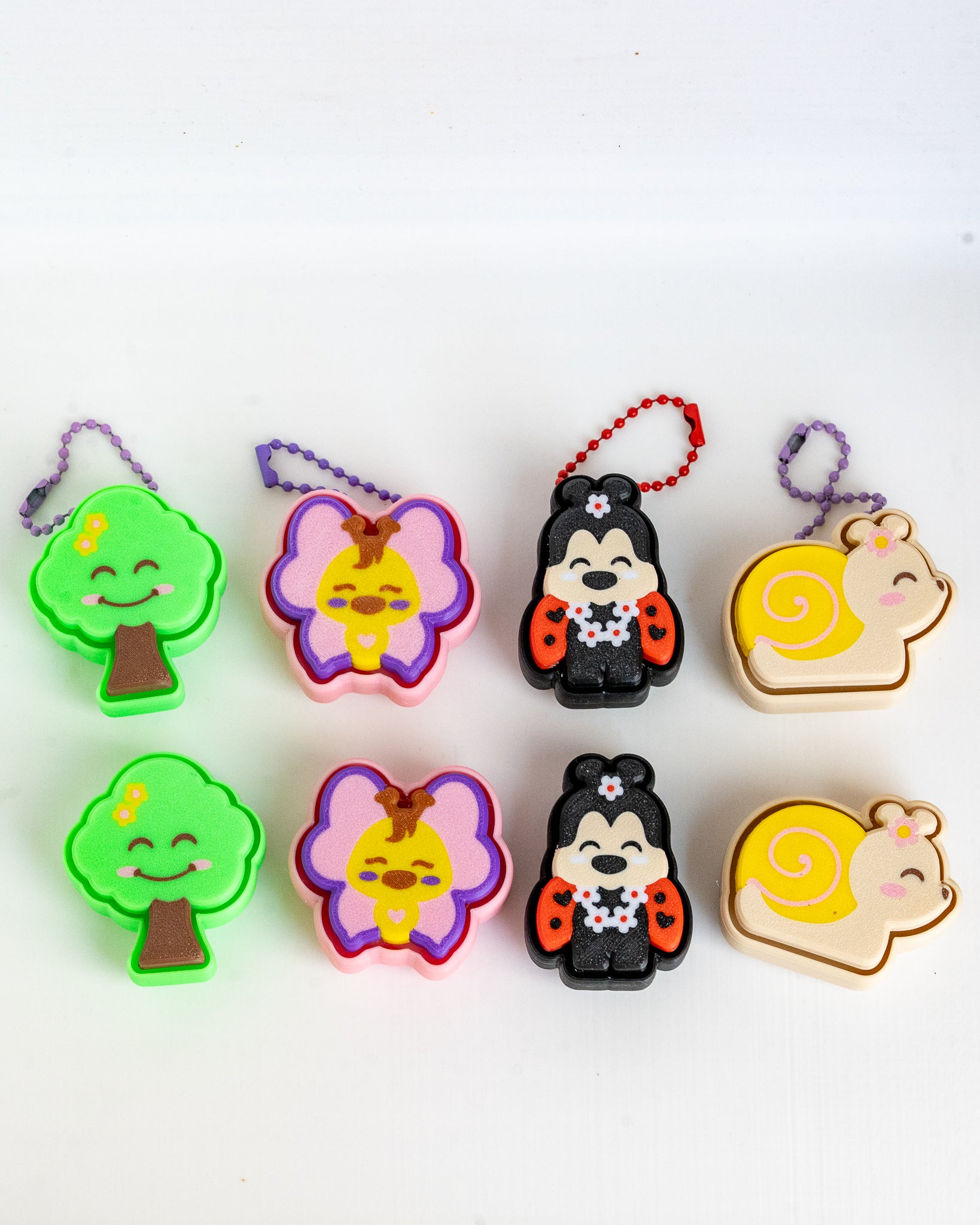 Six small cartoon-shaped keychains rranged in two rows on a plain white background. The set includes tree, butterfly, ladybug, and snail designs in bright colors with hanging loops.