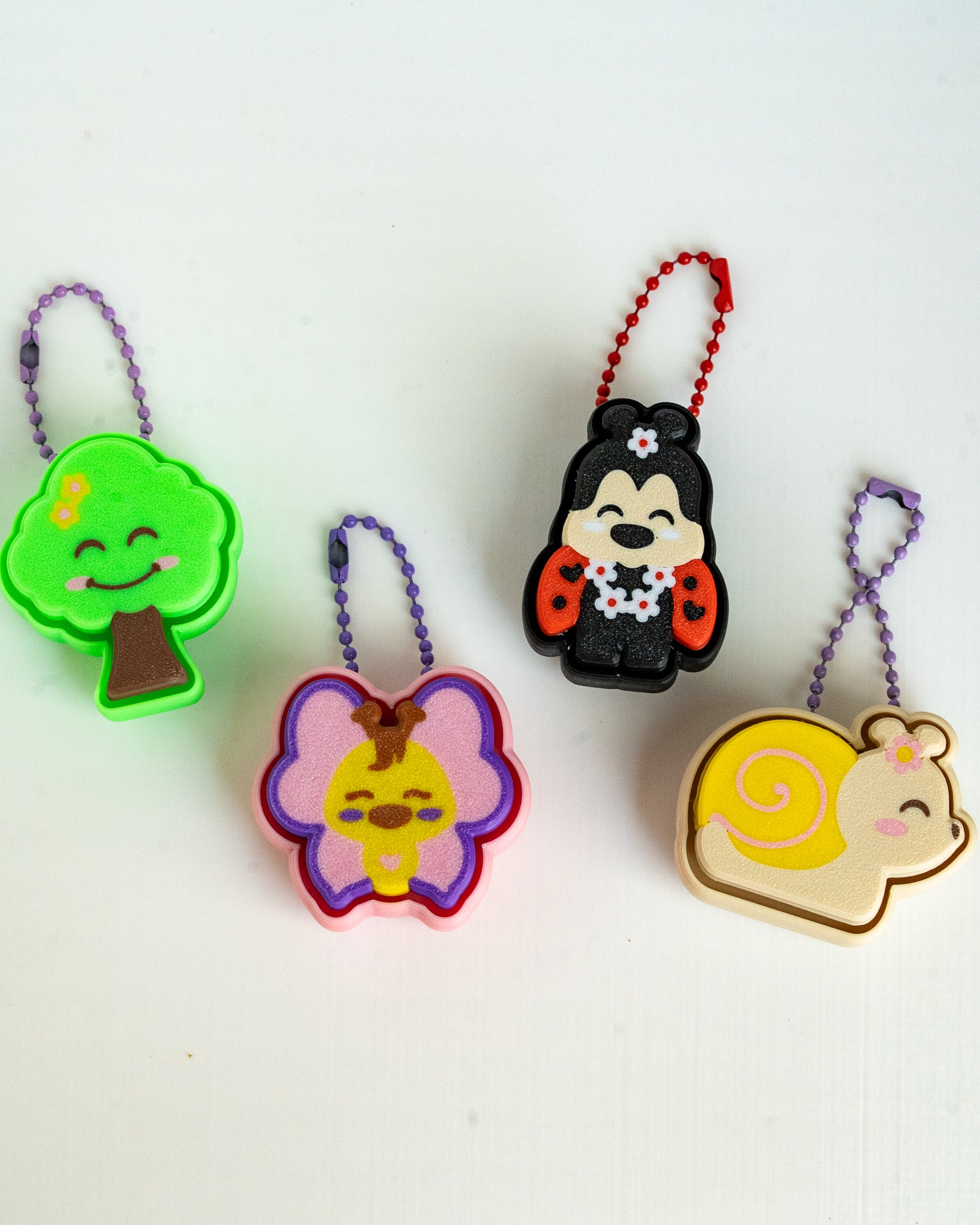 Four small colorful character-shaped keychain charms on a plain white background. The set includes a green tree with a purple chain, a butterfly with a purple and pink chain, a ladybug character with a red chain, and a yellow snail with a purple chain.