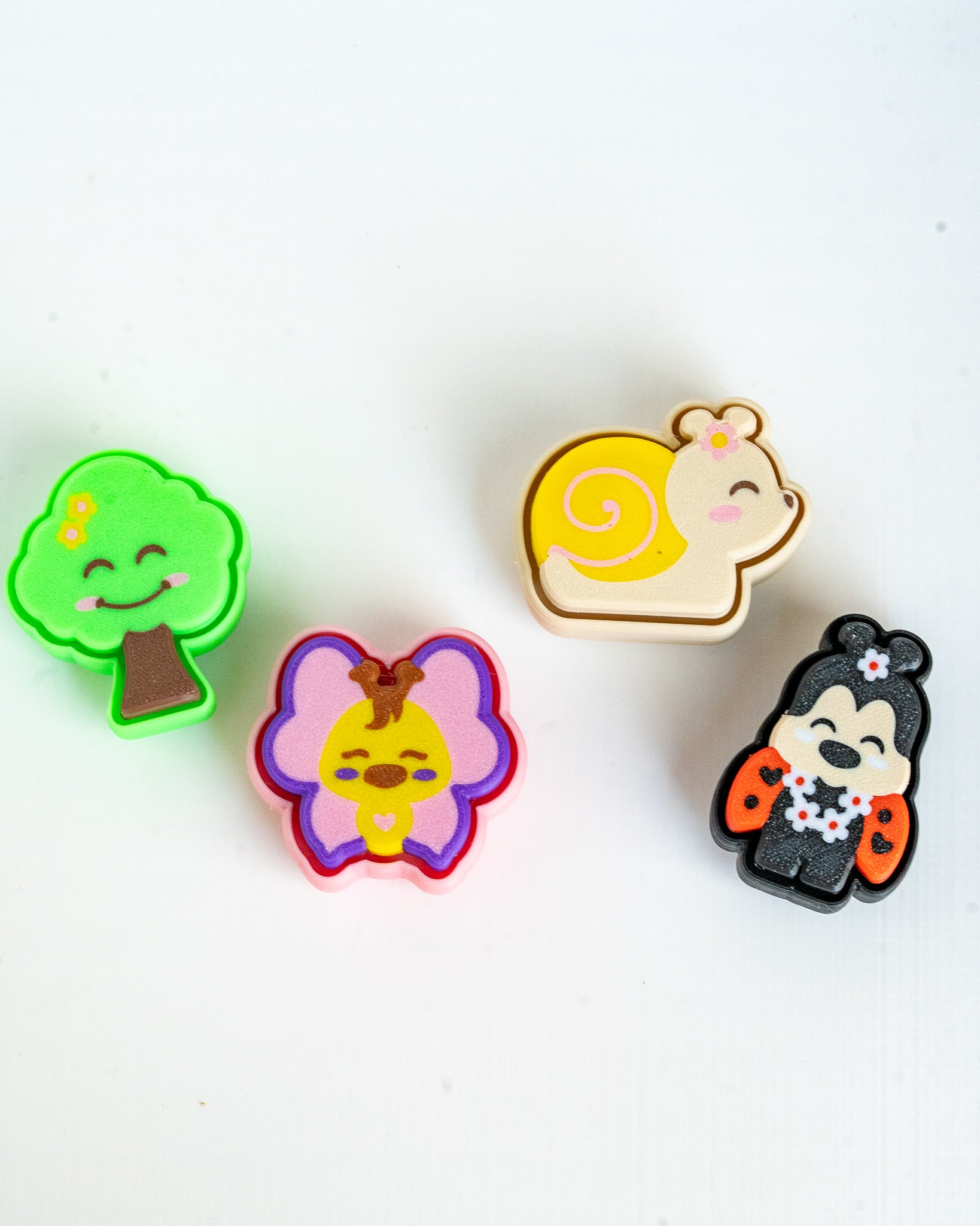 Four small cartoon-shaped clickeers on a plain white background: a green smiling tree, a purple butterfly with a yellow face, a cream snail with a yellow shell, and a ladybug character with red clothing and white flowers.