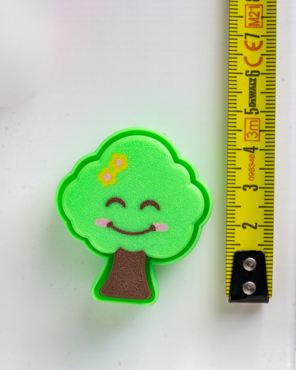 A small green cartoon tree charm or accessory with a smiling face and yellow flowers, shown next to a yellow measuring tape on a white background.