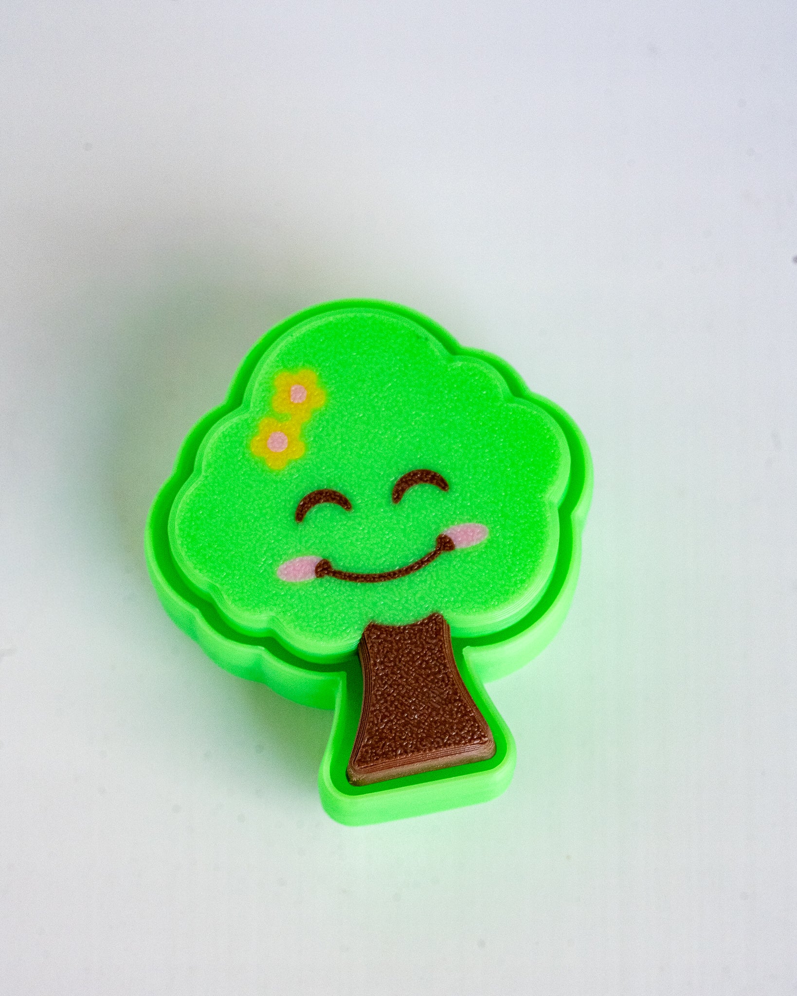 A small tree-shaped decorative item with a bright green body, brown trunk, and a smiling face with pink cheeks and yellow flowers, photographed on a plain white background.