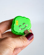 A hand with dark burgundy nail polish holds a small, cloud-shaped green felt pin featuring a smiling face, yellow flowers, and a brown branch against a plain white background.