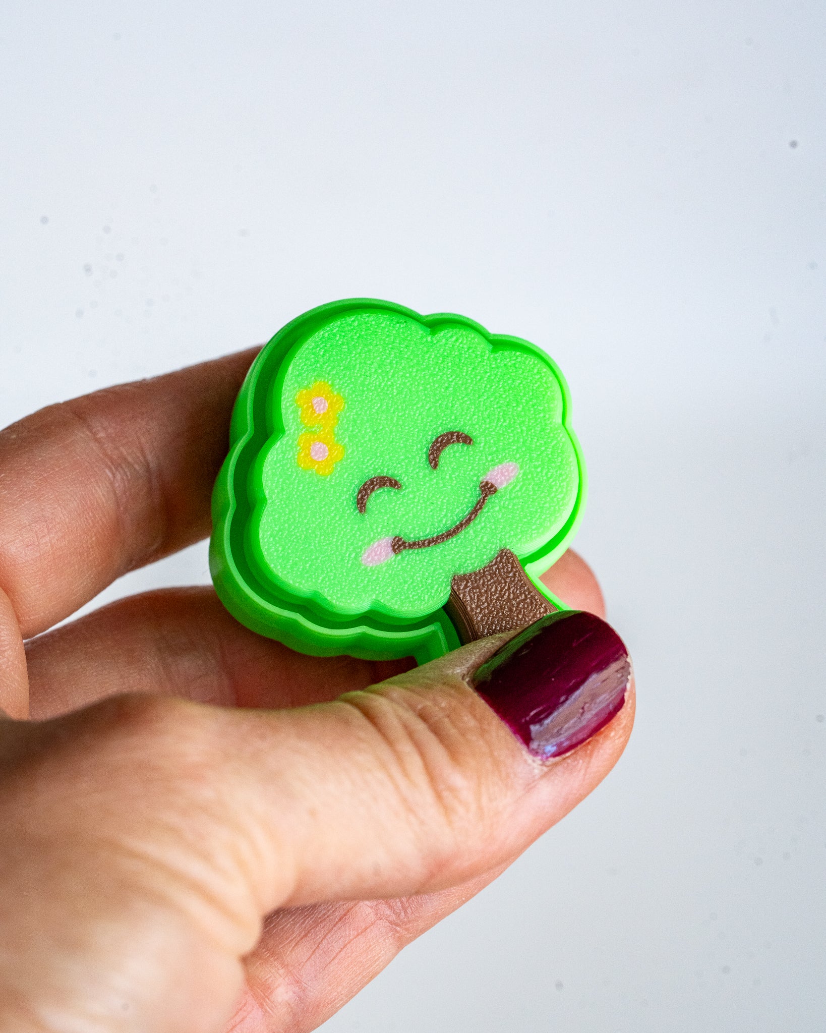 A hand with dark burgundy nail polish holds a small, cloud-shaped green felt pin featuring a smiling face, yellow flowers, and a brown branch against a plain white background.