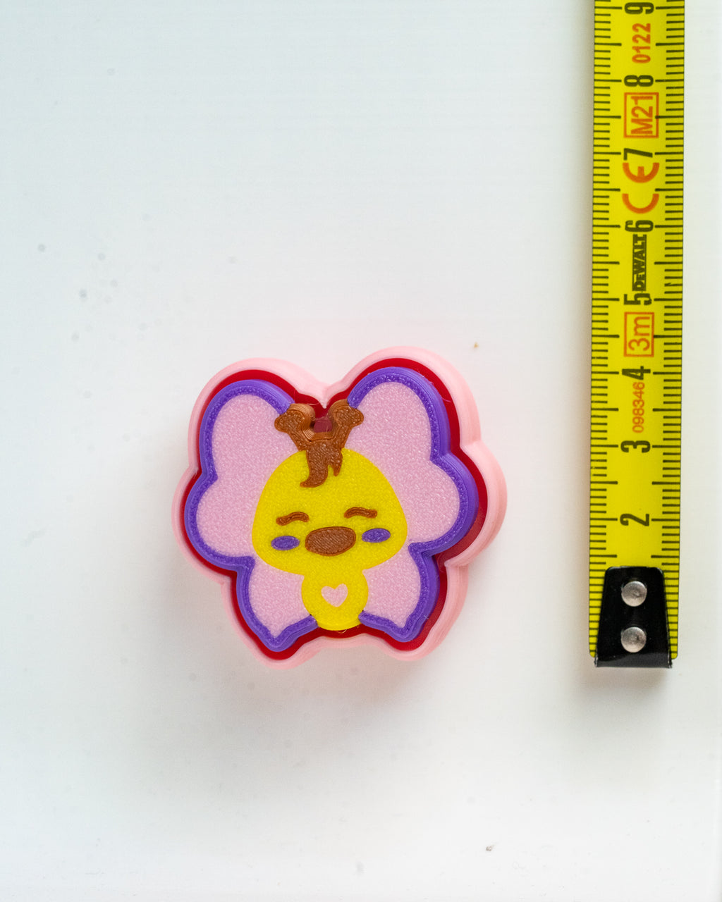 A small pink butterfly-shaped accessory with a cartoon chick design in yellow, purple, and brown, shown next to a yellow tape measure on a white background.