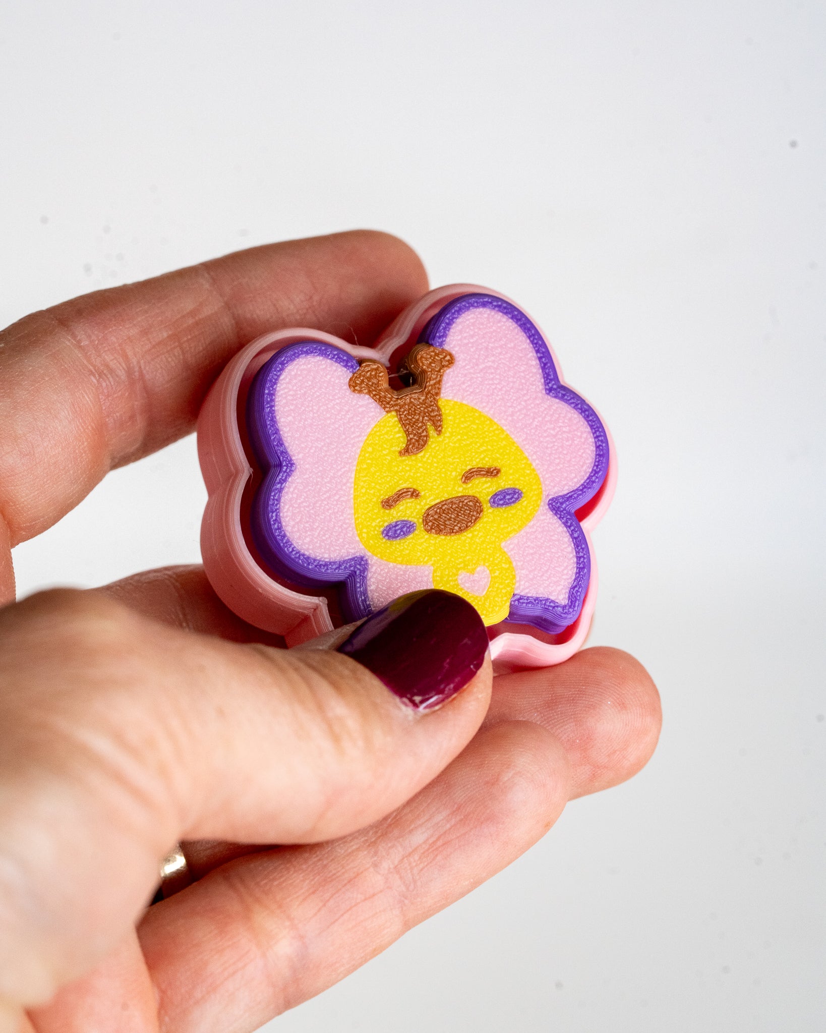 A hand with dark burgundy nail polish holds a small, flower-shaped pink and purple clicker featuring a yellow cartoon chick character on a plain white background.