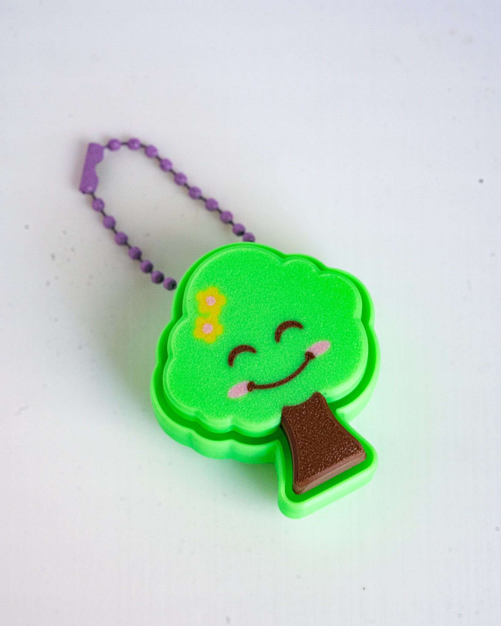 A small tree-shaped keychain or accessory with a bright green body, a smiling face, a yellow flower on the left side, and a brown trunk, attached to a purple ball chain on a white background.