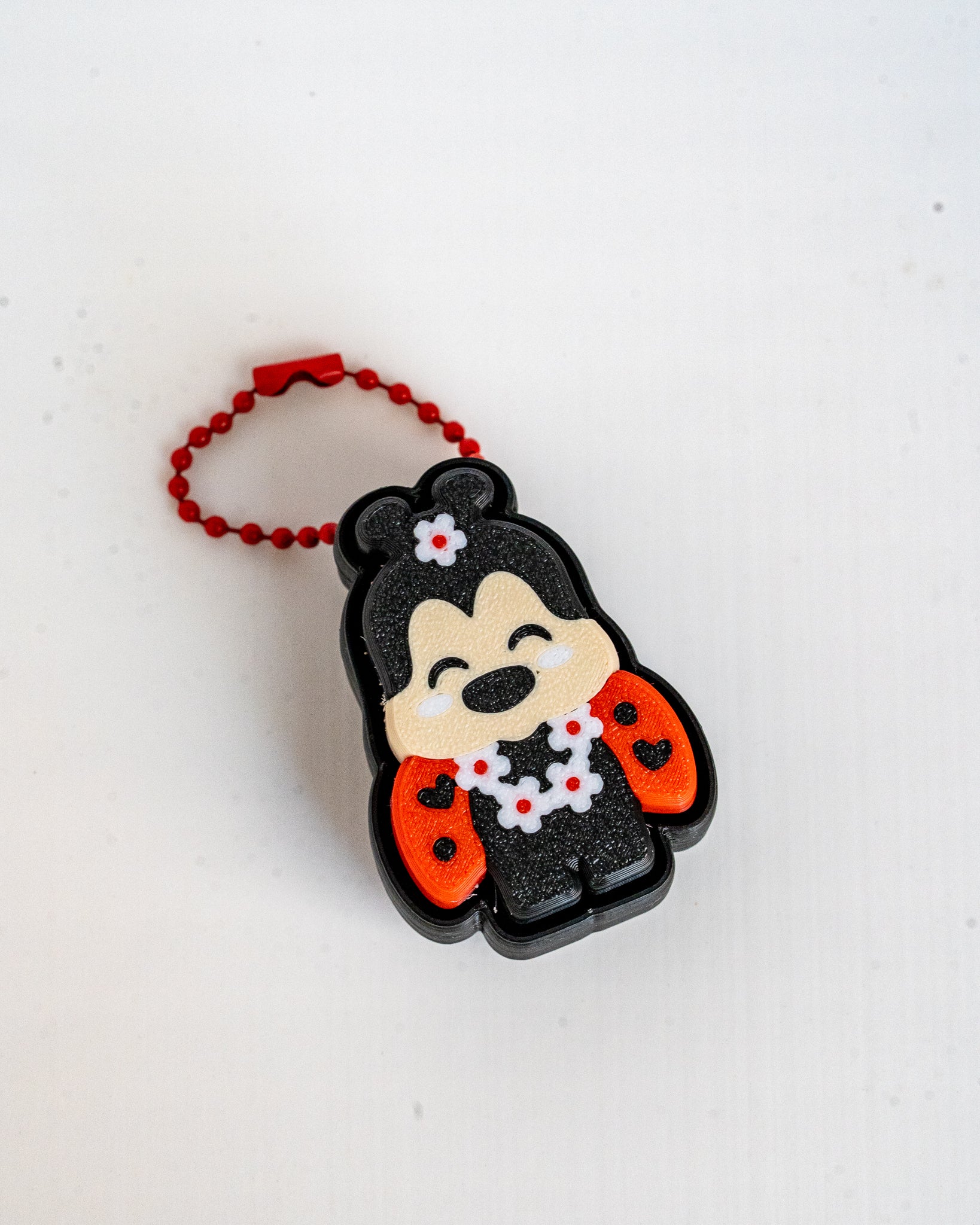 A small lady bug-shaped keychain or bag charm with a red beaded loop, shown on a plain white background. The character is depicted in a black outfit with red sleeves and white flower details.