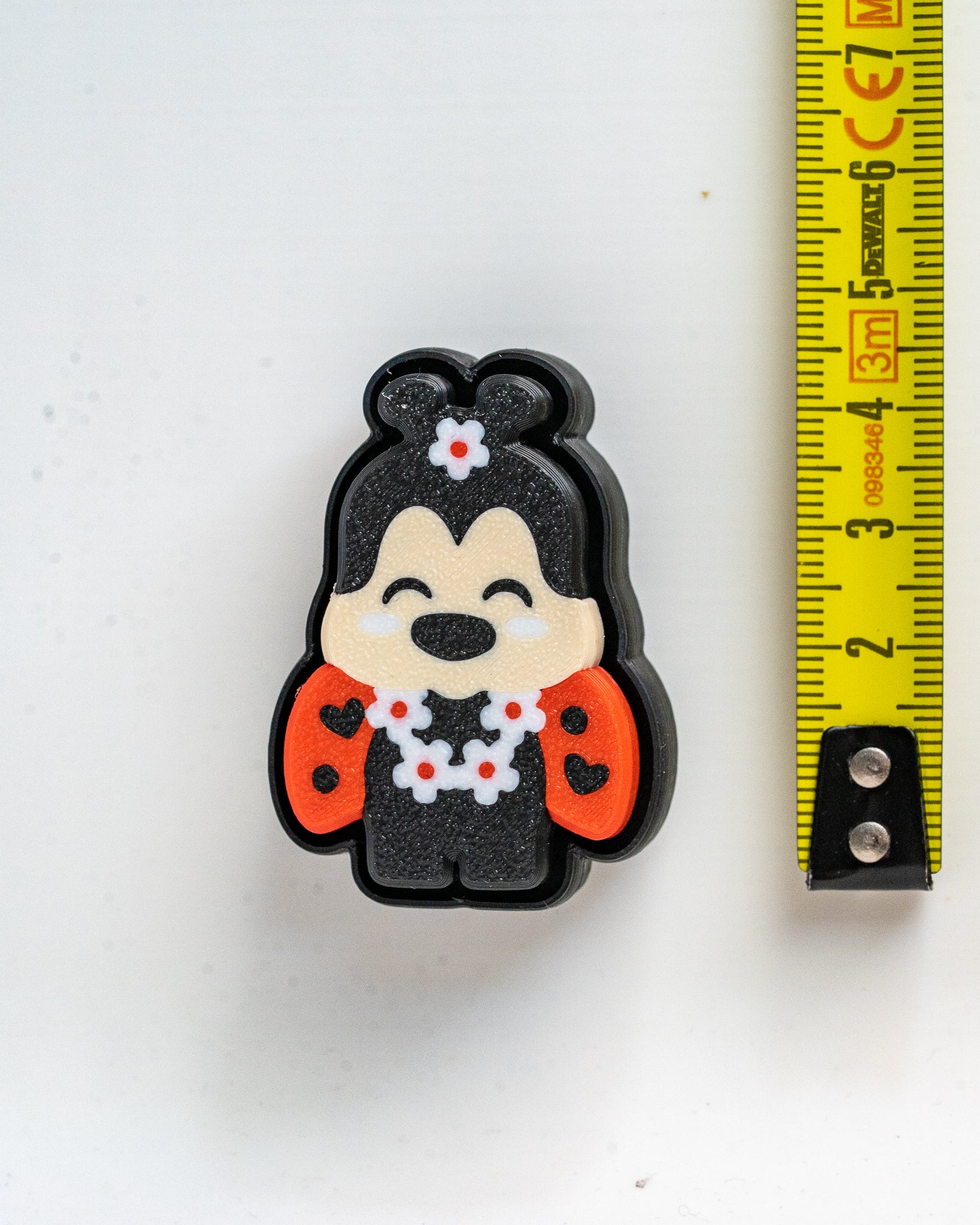 A small black cartoon character charm or accessory with a beige face, red heart-patterned sides, and a white flower lei, shown next to a yellow tape measure on a white surface.