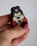 A hand with dark burgundy nail polish holds a small, black and red cartoon character charm against a plain light gray background.