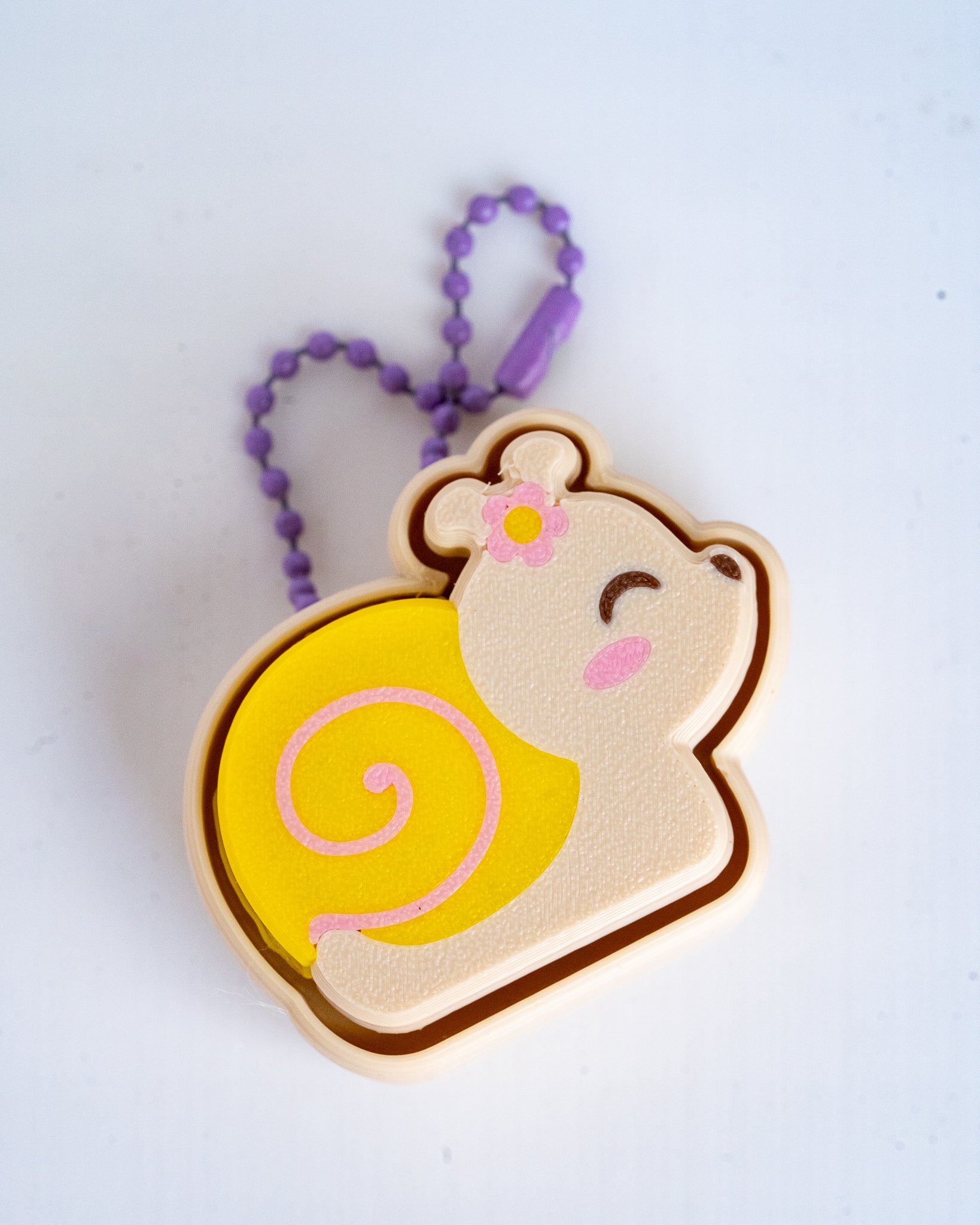 A plastic snail keychain charm with a purple bead chain, featuring a smiling snail with a yellow shell, pink details, and a small flower on its head, set against a plain white background.