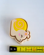 A small plastic figurine shaped like a sleeping cartoon snail, shown on a white background with a yellow tape measure below for scale. The snail has a yellow shell with a pink spiral, a cream-colored body, pink cheeks, and a small pink flower on its ear.