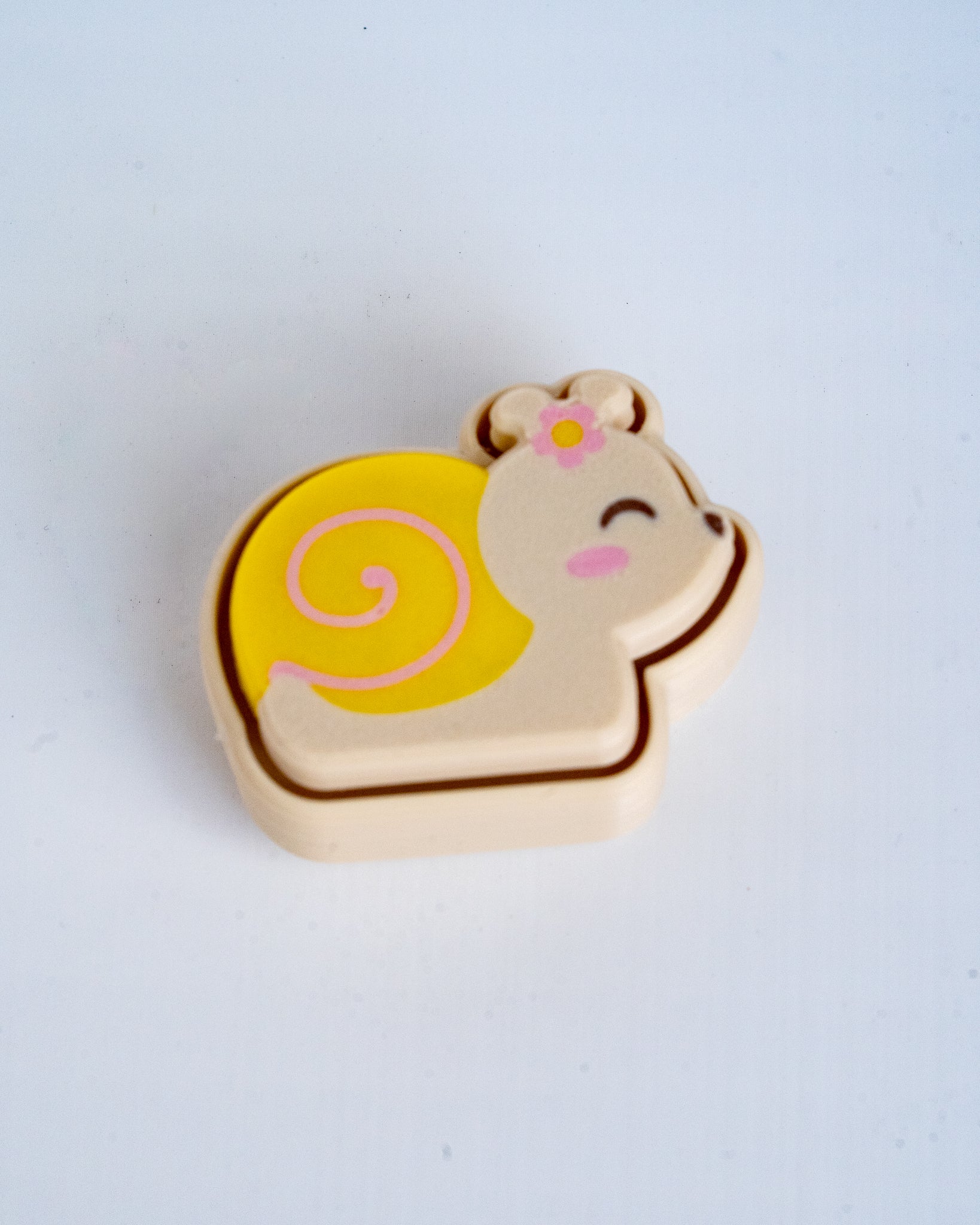 A small plastic toy accessory shaped like a smiling snail, featuring a yellow shell with a pink spiral, a beige body, and a small pink flower on its head, set against a plain light background.