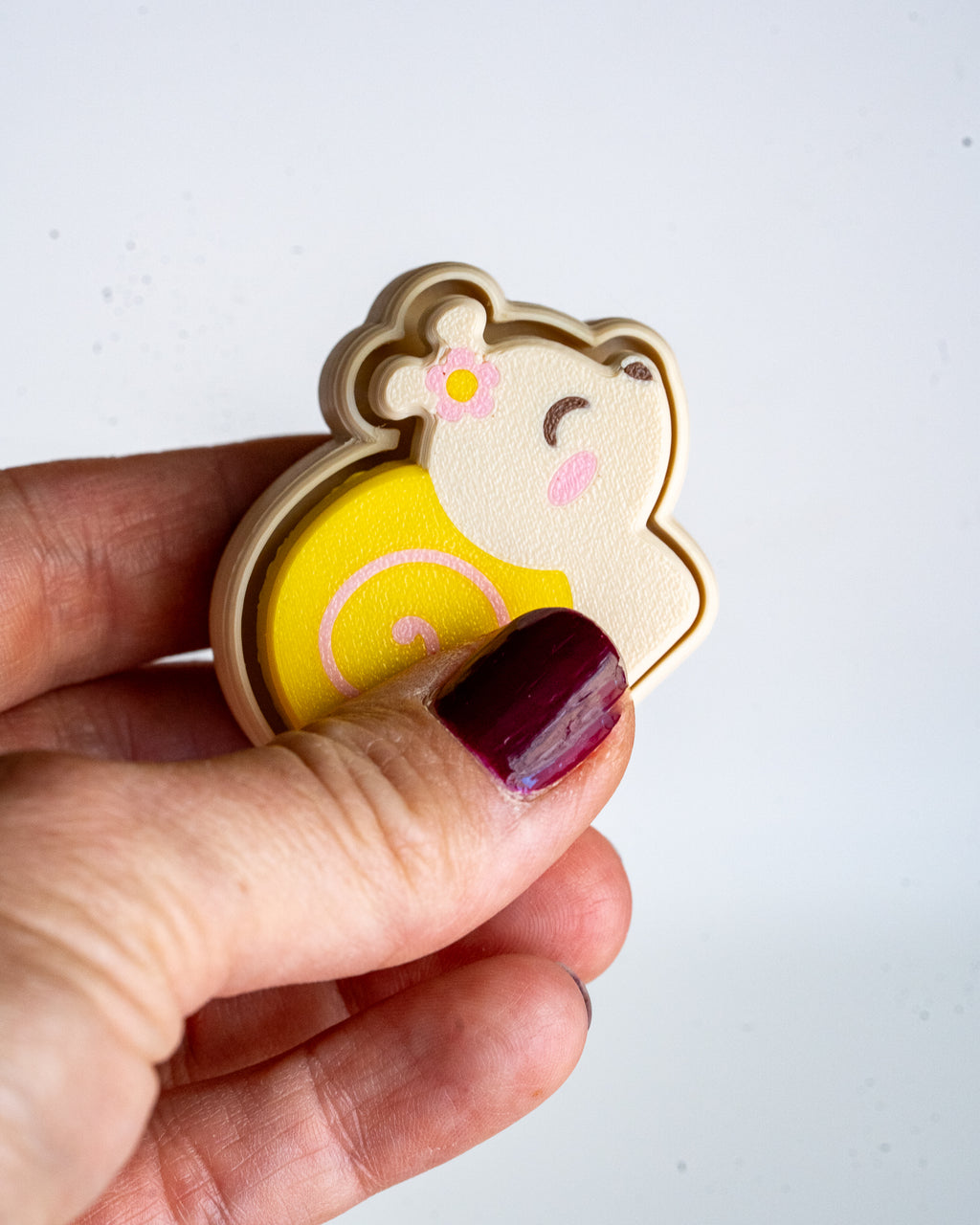 A hand with dark burgundy nail polish holds a small plastic toy shaped like a snail, against a plain white background.