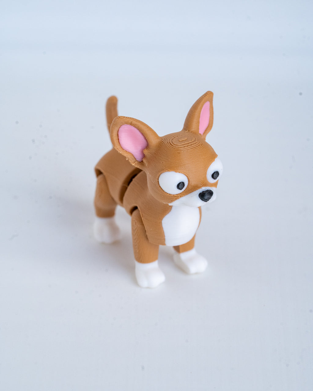 A small plastic toy dog figurine with a brown body, white paws and chest, and pink inner ears, standing on a plain light background.