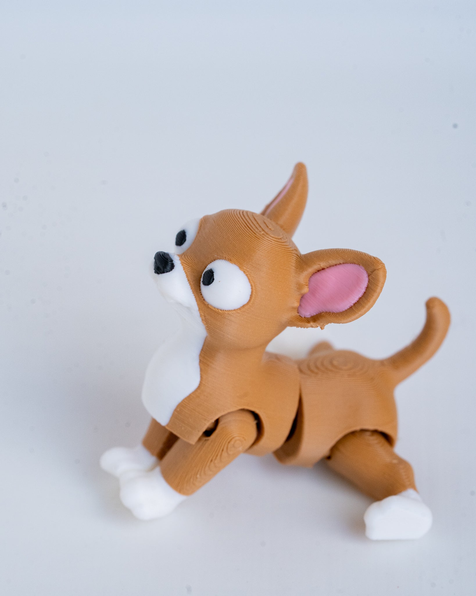 A small articulated dog figurine in a crouching pose, made from brown and white material with pink inner ears and black-and-white eyes, photographed on a plain light background.