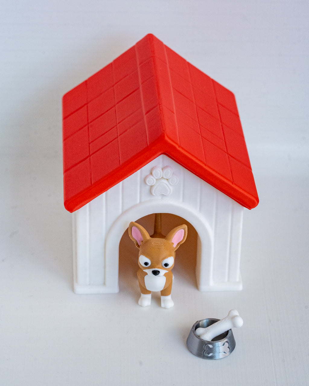 A small plastic dog house toy with a red roof and white body, featuring a tiny tan dog figurine standing in the doorway and a small silver bowl with a white bone in front, all on a plain light background.