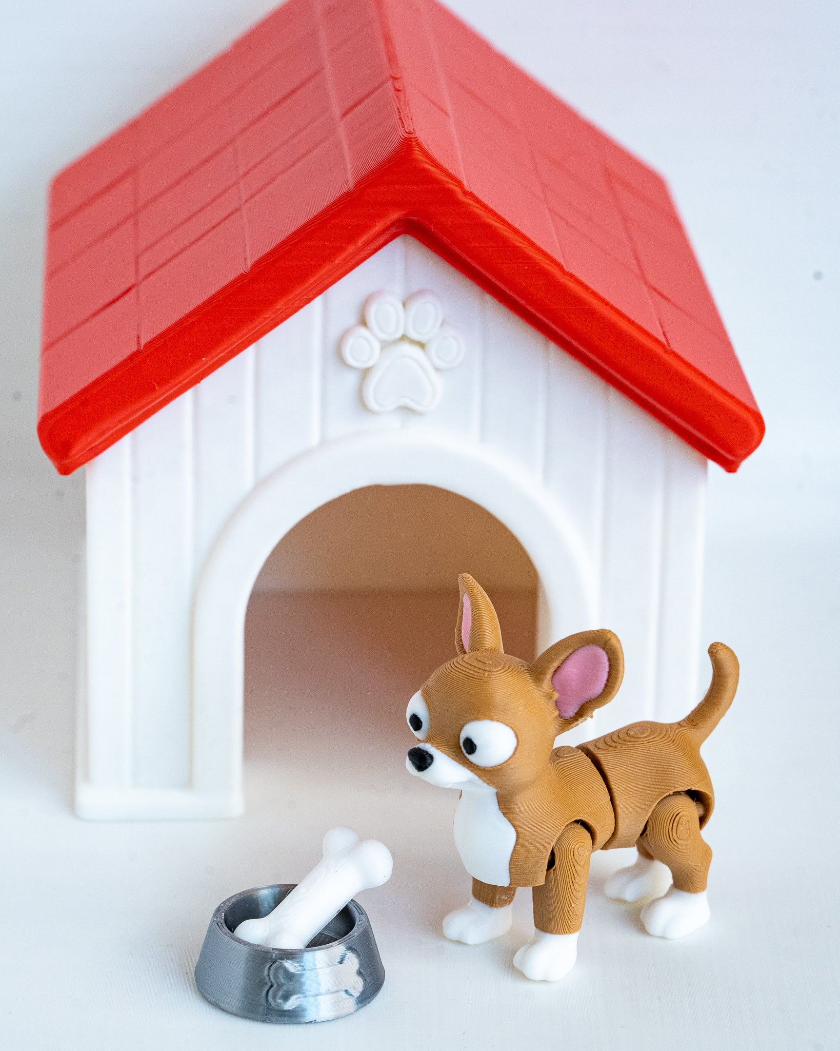 A small brown and white toy dog stands in front of a white dog house with a red roof, next to a silver bowl with a white bone, on a plain white background.