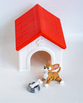 A small plastic dog house toy with a red roof and white body, shown with a brown and white dog figurine and a silver bowl on a plain white background.