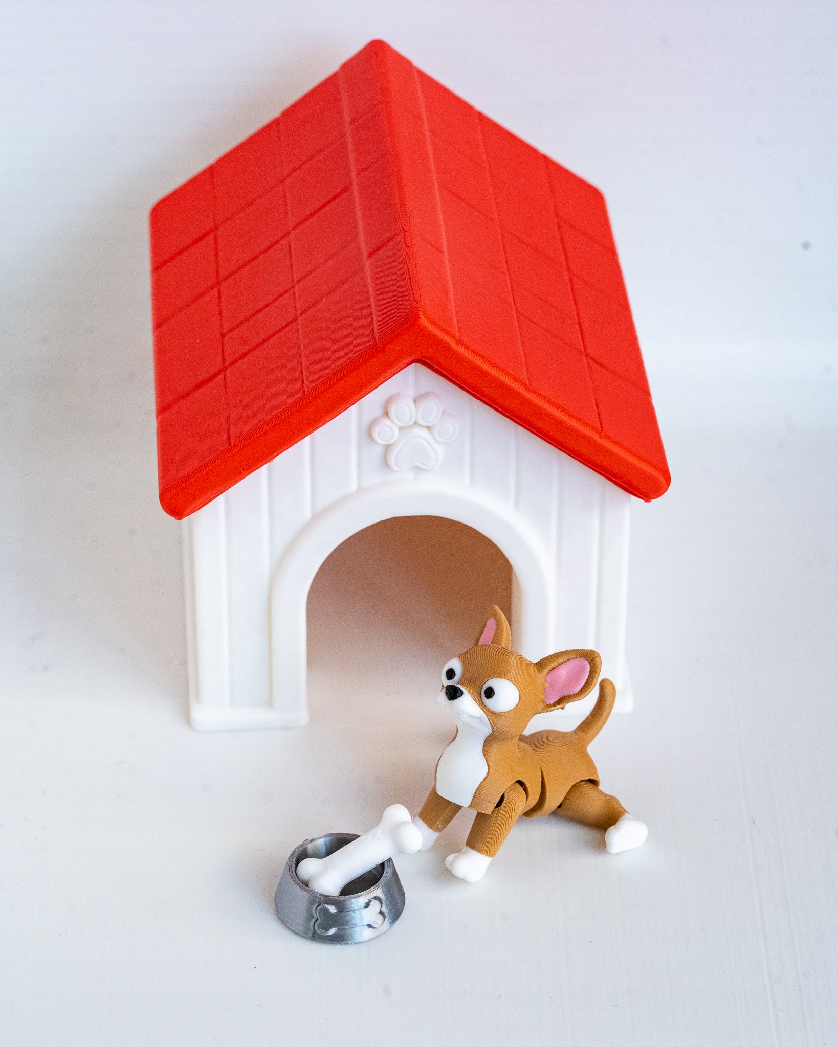 A small plastic dog house toy with a red roof and white body, shown with a brown and white dog figurine and a silver bowl on a plain white background.