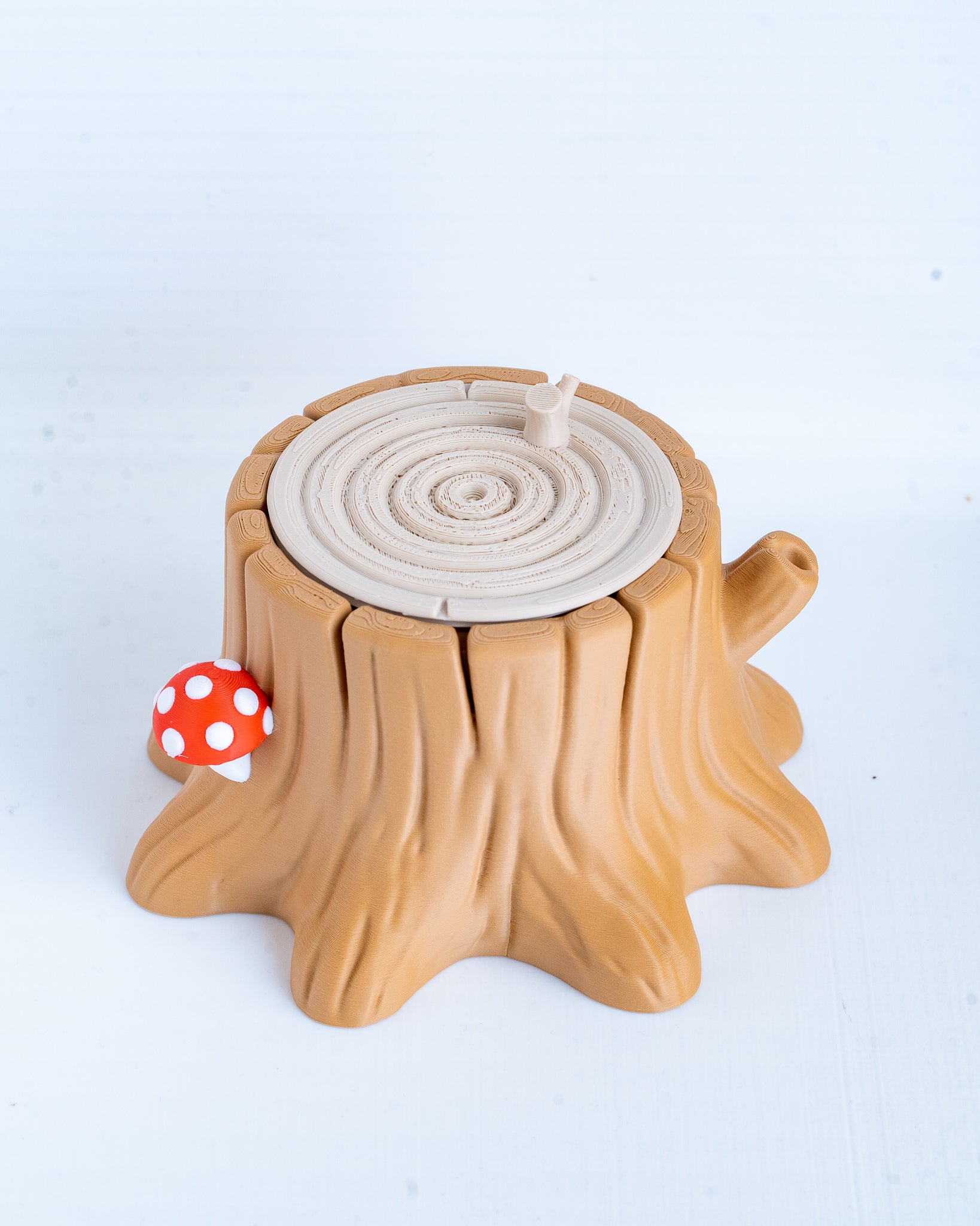 A small toy or decorative stump-shaped object in tan and light brown, with a circular wood-grain pattern on top, a red-and-white polka dot mushroom attached on the left side, photographed on a plain light background.