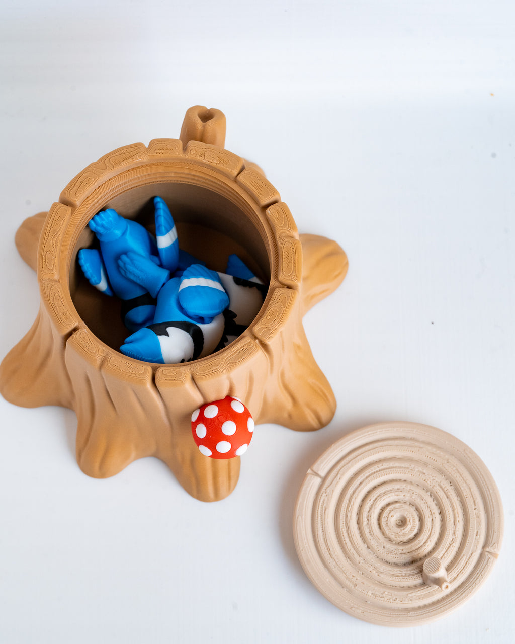 A plastic figurine of a tree stump container with its lid placed beside it on a white background. The stump is light brown and contains two small blue-and-white cartoon fish figures, with a small red mushroom accent on the side