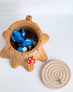 A plastic figurine of a tree stump container with its lid placed beside it on a white background. The stump is light brown and contains two small blue-and-white cartoon fish figures, with a small red mushroom accent on the side
