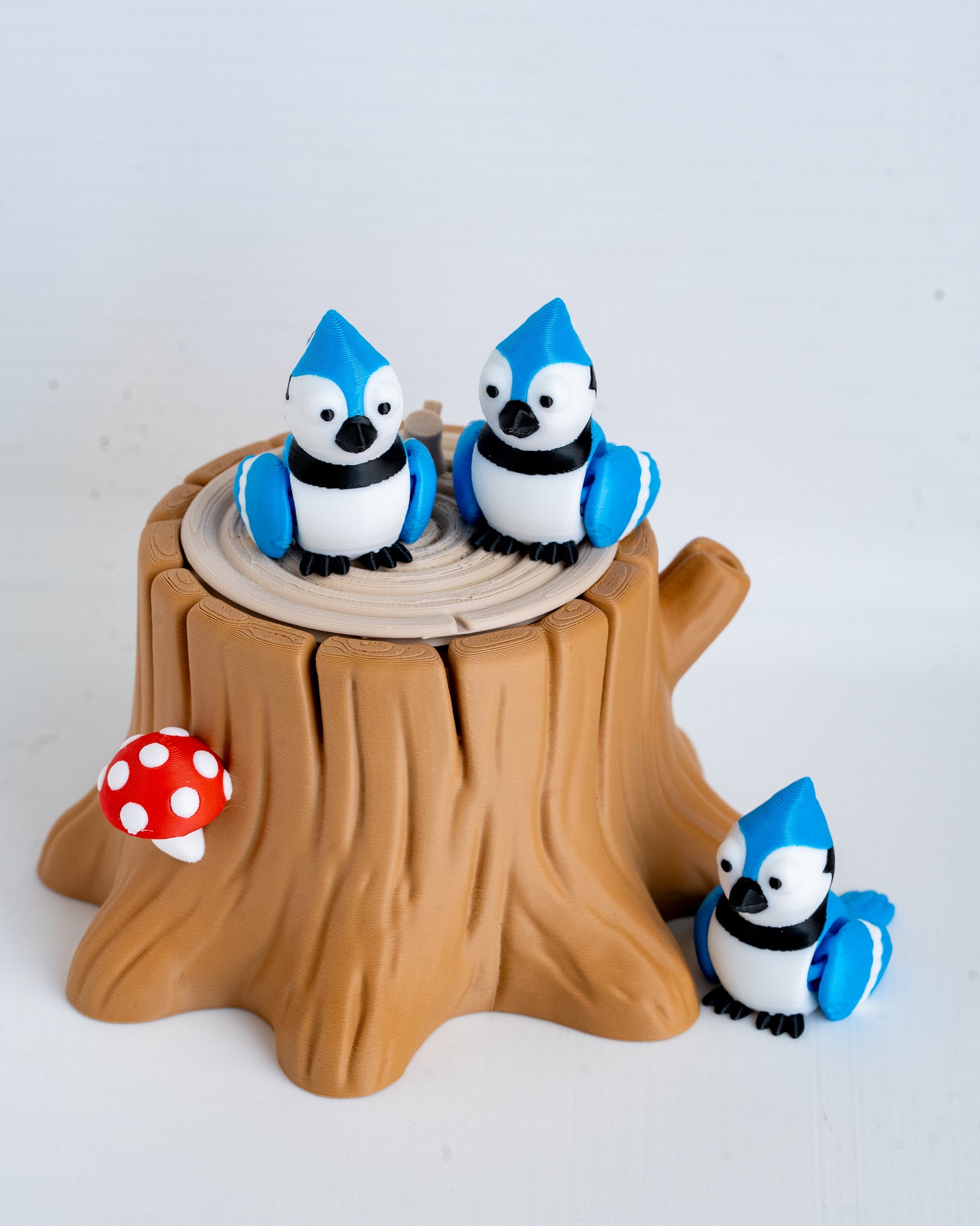 A set of small bluebird figurines posed around a brown tree stump display, with one bird on each stump top and one on the side, plus a red-and-white mushroom accent, photographed on a clean white background.