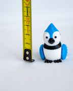 A small blue, white, and black blue jay figurine standing next to a yellow tape measure on a plain white background.