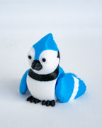 A small blue jay figurine with a bright blue and white body, black beak and feet, and a black band around the neck, photographed on a plain light background.
