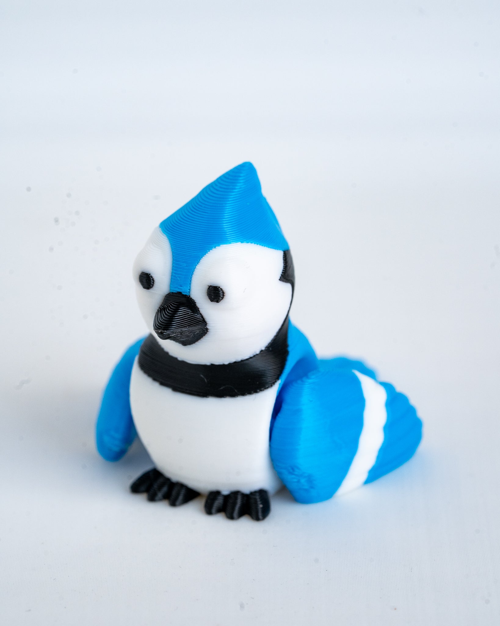 A small blue jay figurine with a bright blue and white body, black beak and feet, and a black band around the neck, photographed on a plain light background.
