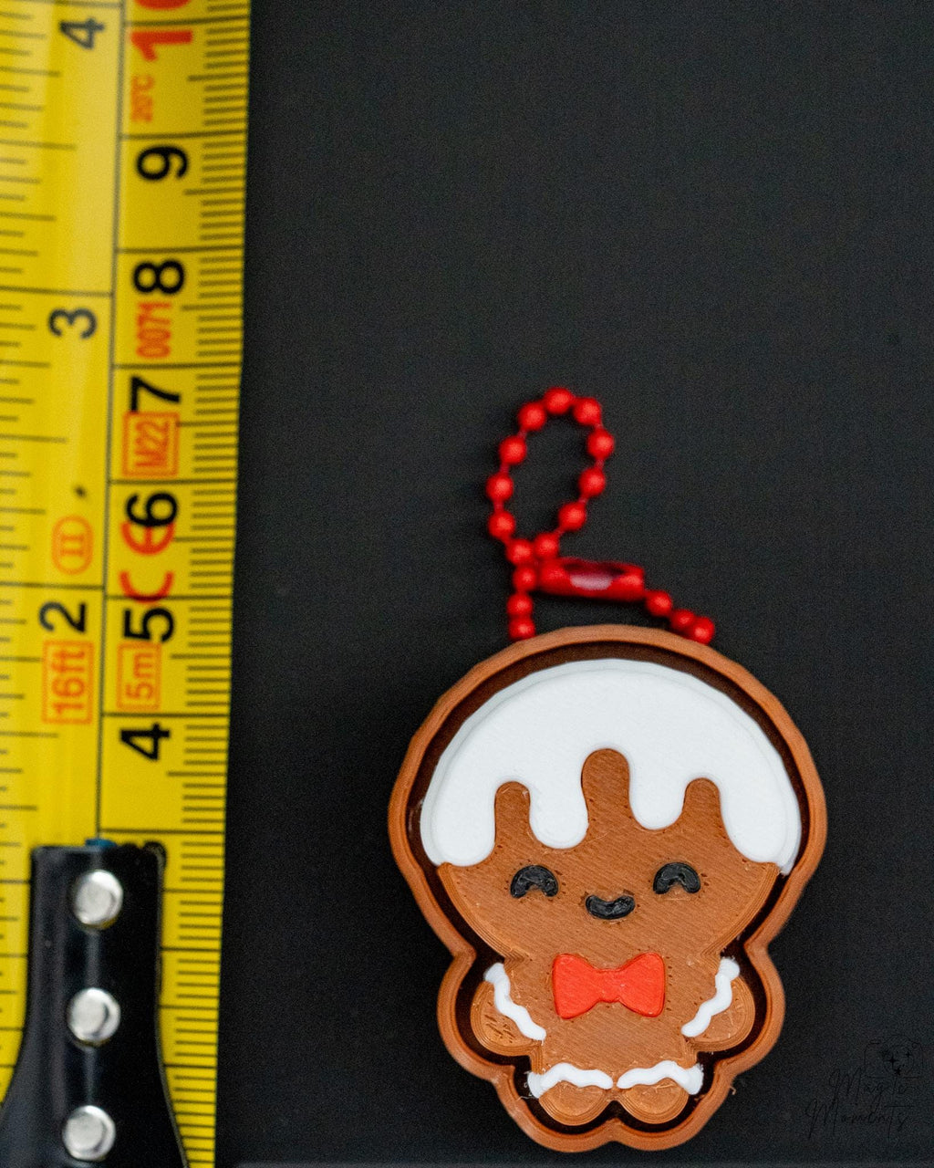 Gingerbread man keychain with a red bow tie next to a ruler on a black background