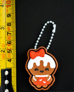 Gingerbread girl keychain with a red bow and white icing on a black background, next to a ruler for scale.