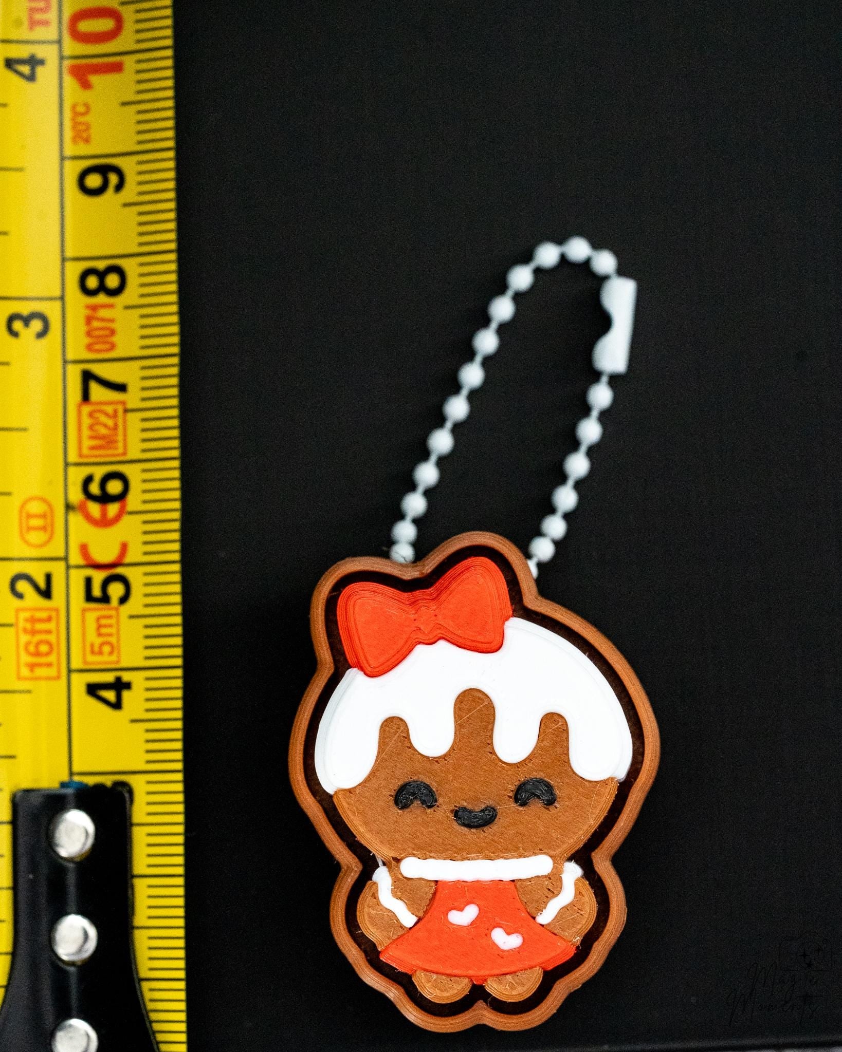 Gingerbread girl keychain with a red bow and white icing on a black background, next to a ruler for scale.